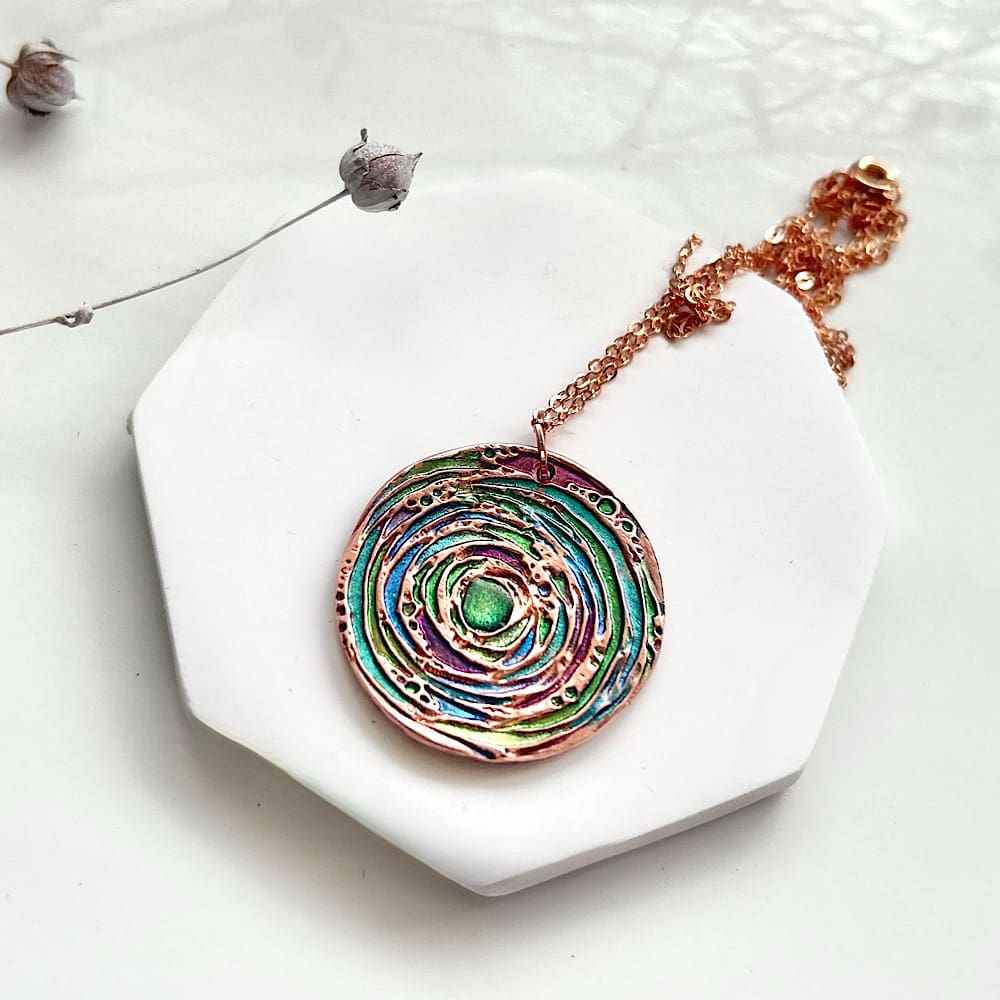 A round handmade copper pendant that has been hand painted with bright metallic colours.The necklace has a rose gold plated chain and the image has a White background.