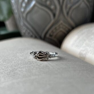 A small delicate ring with a floral bud pattern lies on top of an open book. you can see a grey flower pot in the background