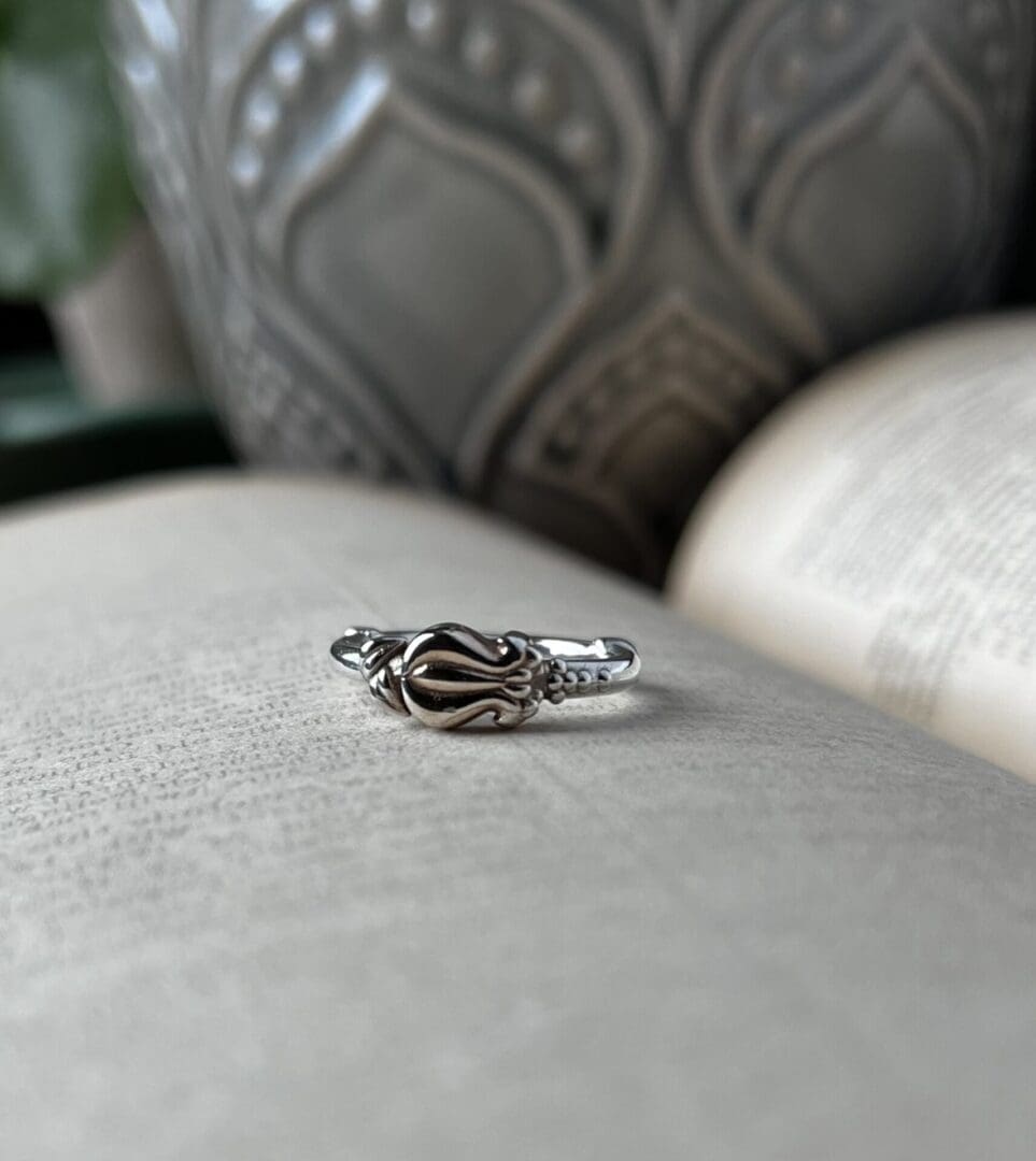 A small delicate ring with a floral bud pattern lies on top of an open book. you can see a grey flower pot in the background