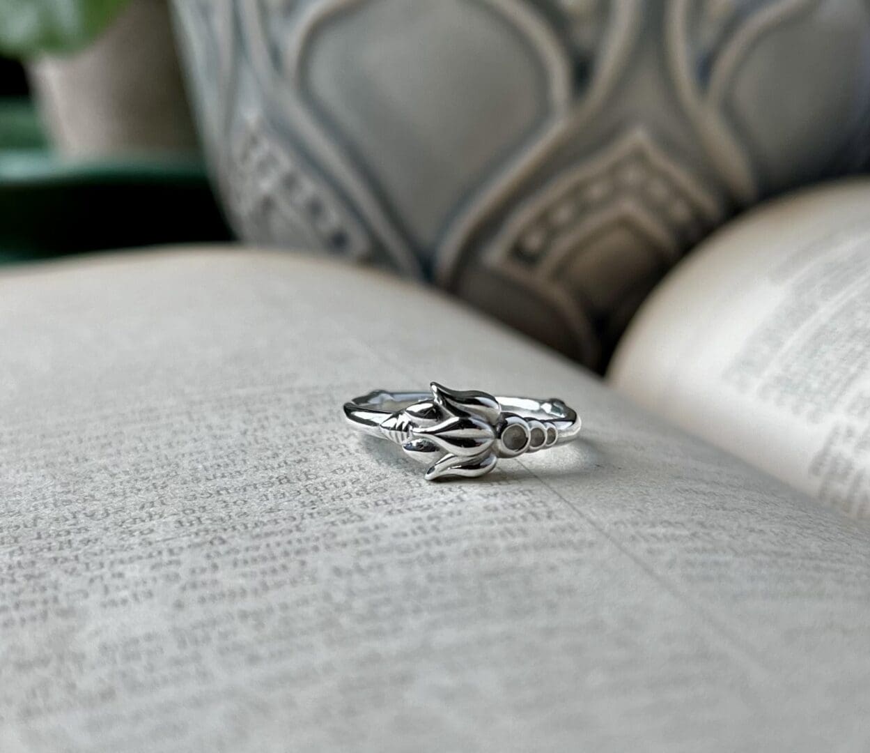 a delicate silver ring with a flower-style pattern on it lies on top of an open book. You can see a grey flower pot in the background.