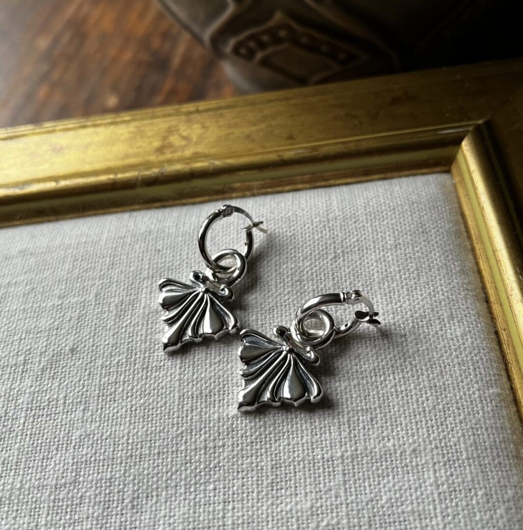 a pair of silver earrings with an art deco pattern and hoop fittings. They lie on top of a natural white fabric, you can see a gilded worn out frame on the background.