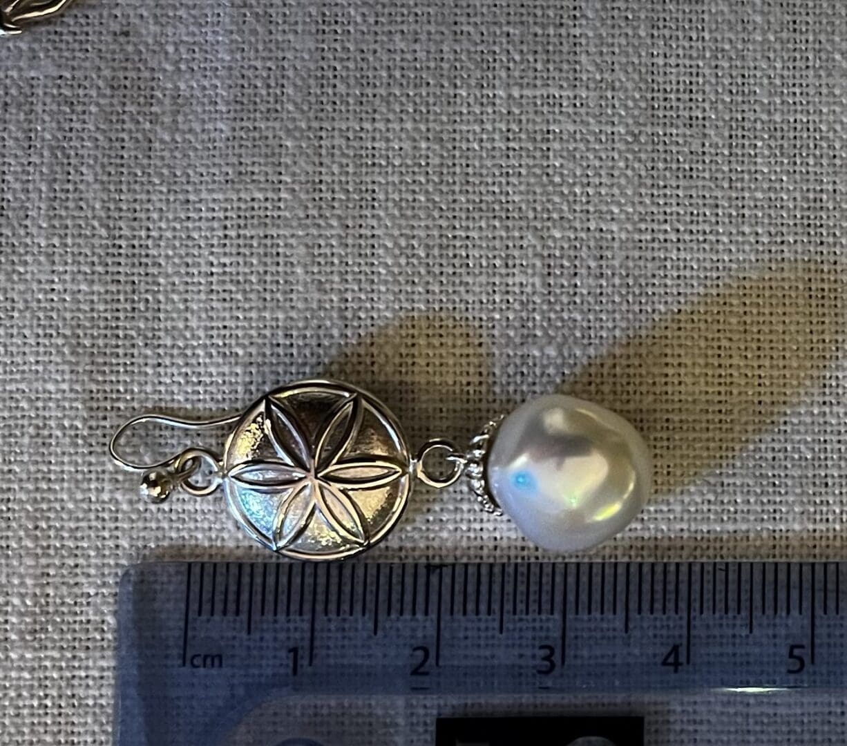 An earring with a silver daisy wheel symbol and a baroque pearl underneath it. There is a ruler next to the earring stating the total drop length, it says 36mm.