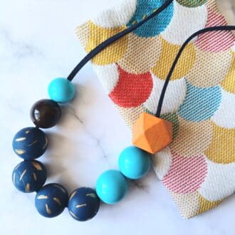 A chunky colourful beaded necklace is on a white marble background. Part of the necklace is resting on a fabric pouch made from multicoloured scallop pattern fabric. The necklace has a mixture of light blue vintage plastic beads, painted mid blue wooden beads with a gold sprinkle pattern and one orange larger wooden geometric bead. The beads are threaded on a length of navy blue faux cord fastened with an adjustable sliding knot.