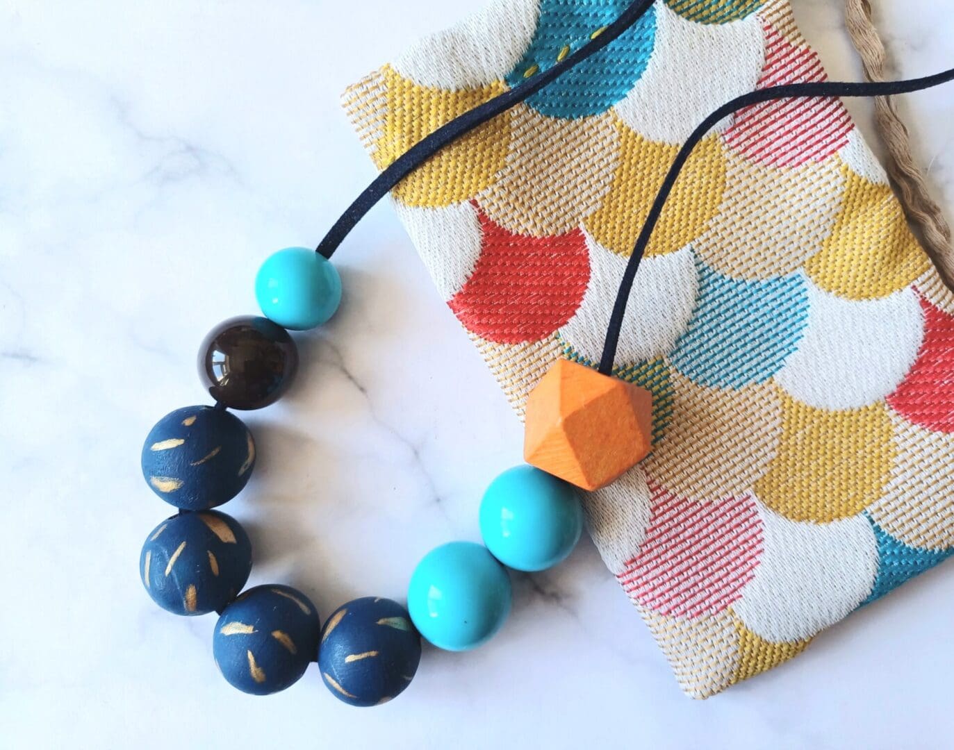 A chunky colourful beaded necklace is on a white marble background. Part of the necklace is resting on a fabric pouch made from multicoloured scallop pattern fabric. The necklace has a mixture of light blue vintage plastic beads, painted mid blue wooden beads with a gold sprinkle pattern and one orange larger wooden geometric bead. The beads are threaded on a length of navy blue faux cord fastened with an adjustable sliding knot.