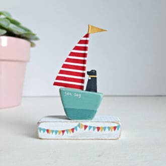 Tiny wooden sailing boat with a sea green base and a red and white stripe sail and yellow flag. A tiny black dog is sitting in the boat