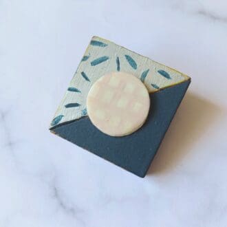 A square brooch made of wood with a diagonal line separating the two halves. One triangular half is painted navy blue, the other is painted silver with a pattern of short navy blue lines in a random pattern. In the centre of the square is a white circle painted with a cream grid pattern. The brooch is on a white and grey marble patterned background.