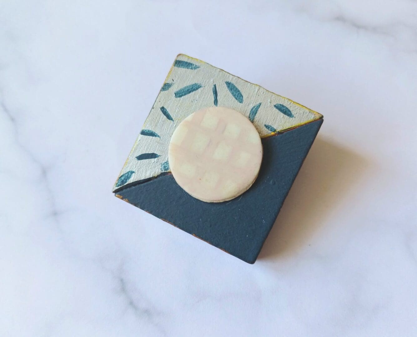 A square brooch made of wood with a diagonal line separating the two halves. One triangular half is painted navy blue, the other is painted silver with a pattern of short navy blue lines in a random pattern. In the centre of the square is a white circle painted with a cream grid pattern. The brooch is on a white and grey marble patterned background.