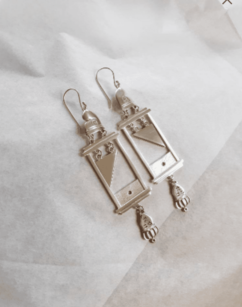 A pair of guillotine earrings in silver. There is a phrygian cap on top of the frame, a silver blade is suspended by small loops and underneath the frame is a head upside down