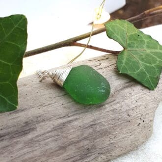 Bright green sea glass with a silver wire wrapped top hanging on a silver chain displayed on a piece of driftwood with ivy leaves