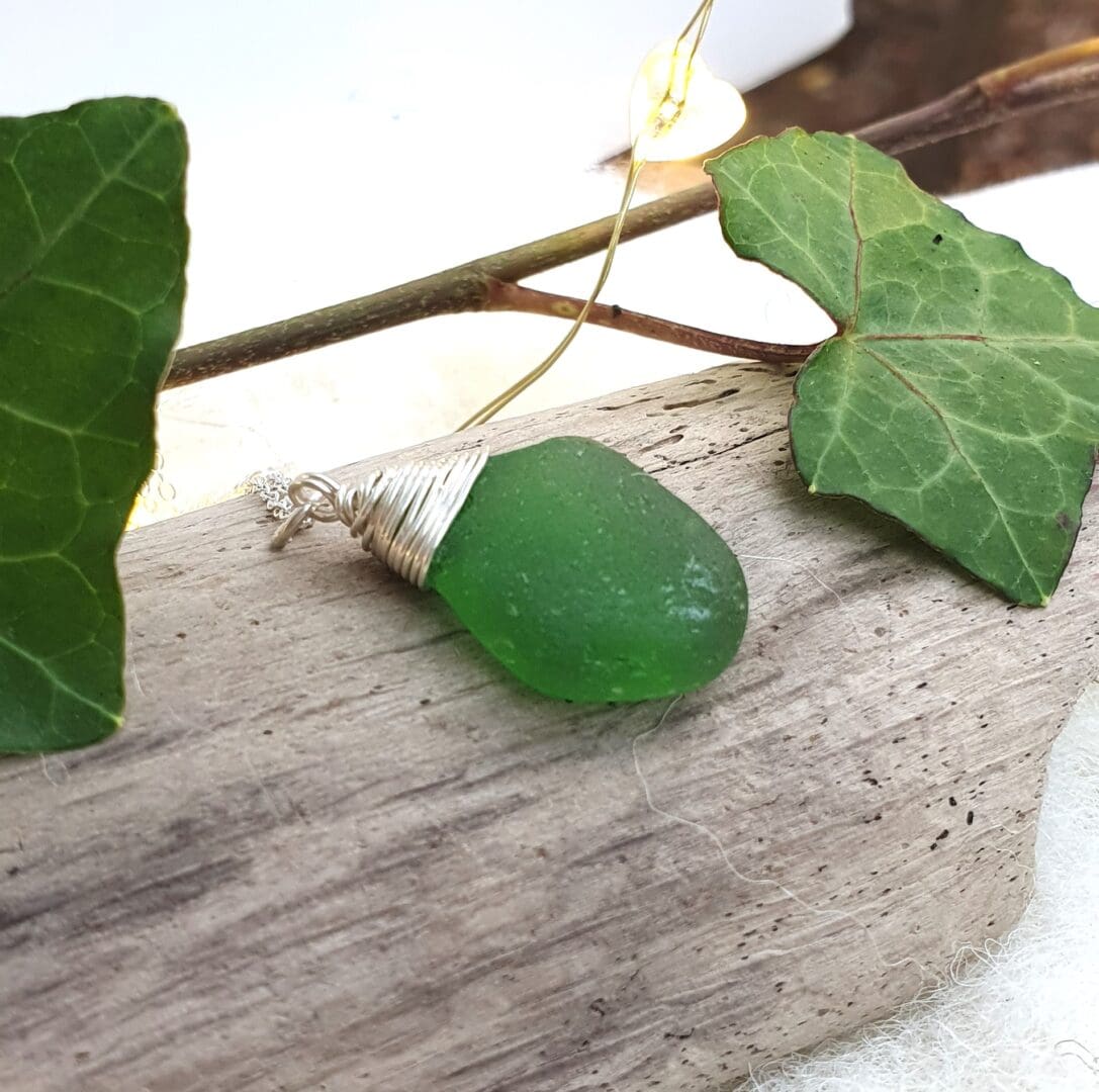 Bright green sea glass with a silver wire wrapped top hanging on a silver chain displayed on a piece of driftwood with ivy leaves