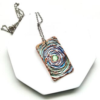 A oblong copper pendant, with a circular pattern and hand-painted in pastel metallic colours. With a fine sterling silver chain.