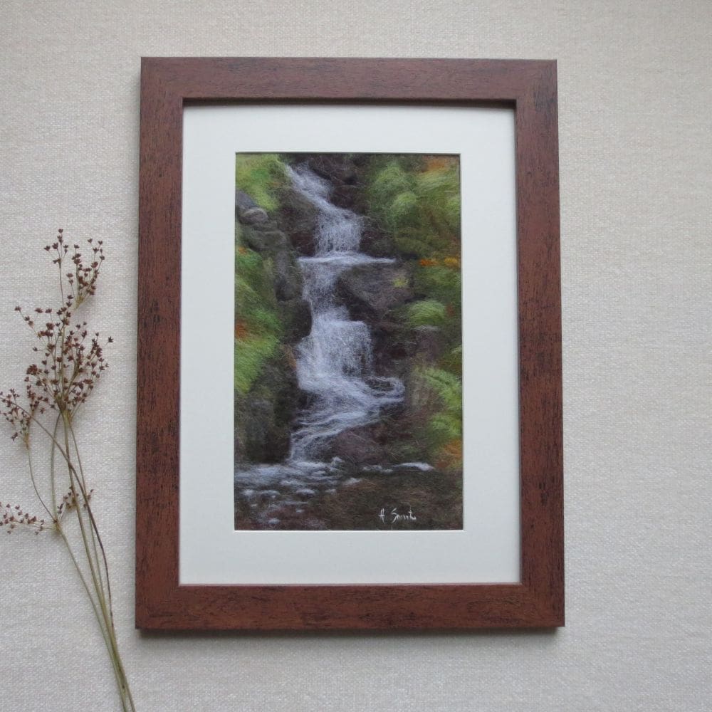 Painting made from wool depicting a small babbling waterfall running down over moss covered stones. Framed in a dark wood effect frame with a cream coloured mount.