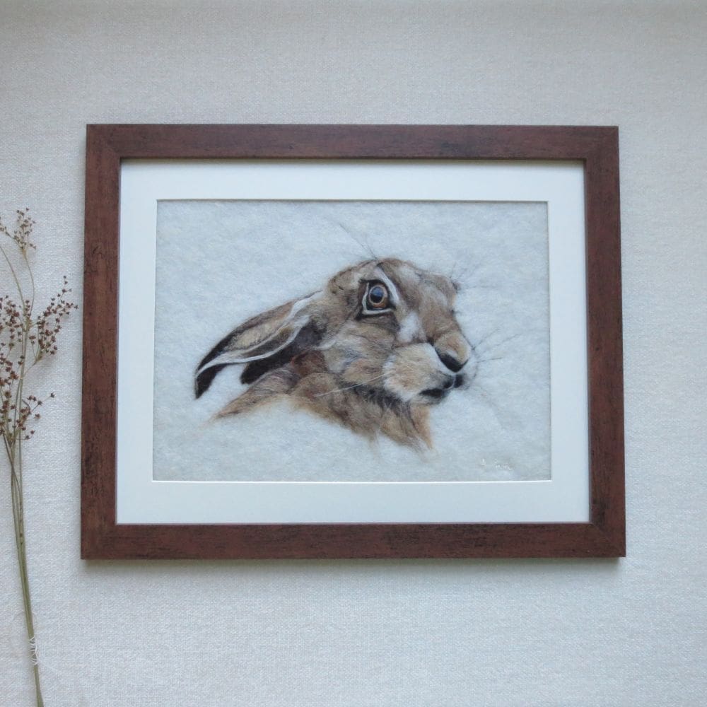 The picture depicts the head and shoulders of a brown hare. The hare is facing forward but looking back over it's right shoulder with it's ears down. The background is cream and the frame is dark brown.