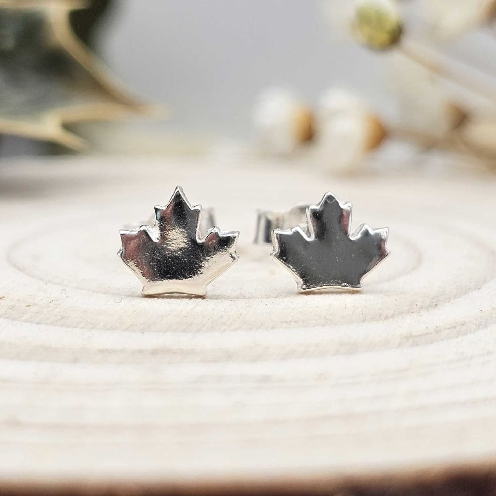 Pair of handcrafted recycled shiny sterling silver maple leaf shaped stud earrings sat on wooden disk