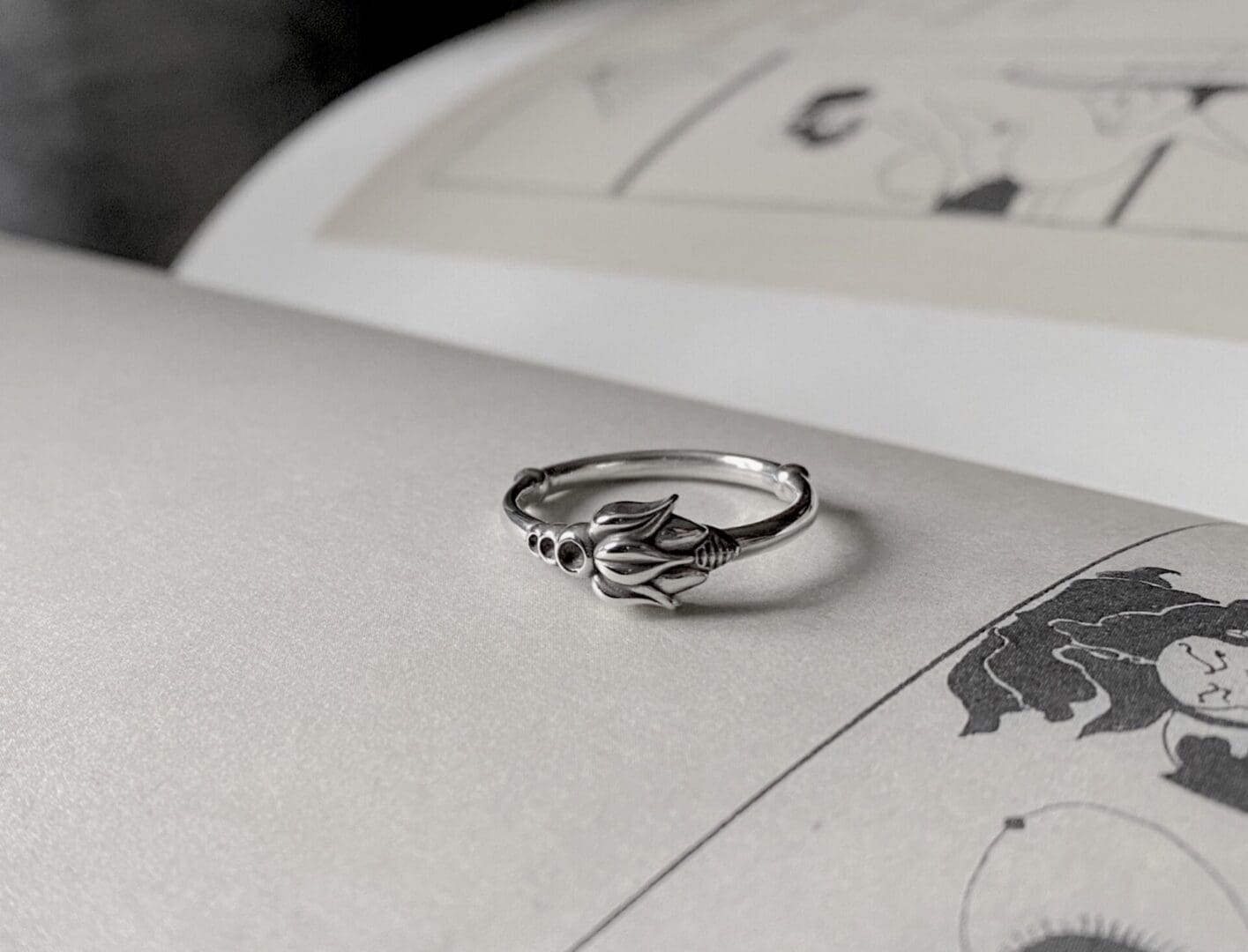 a delicate silver ring with a flower pattern lies on top of an open art book, you can see parts of a print on the pages.