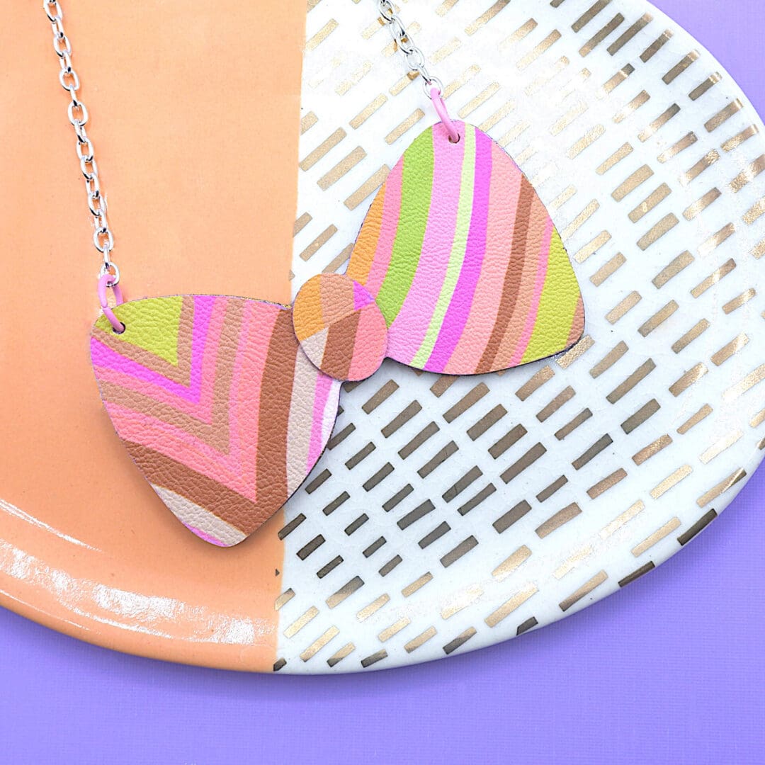 Handmade necklace shaped like a bow tie. Colourful hand draw stipe effect pattern in pinks, oranges with dashes of yellow and lime. Faux leather and painted plywood construction