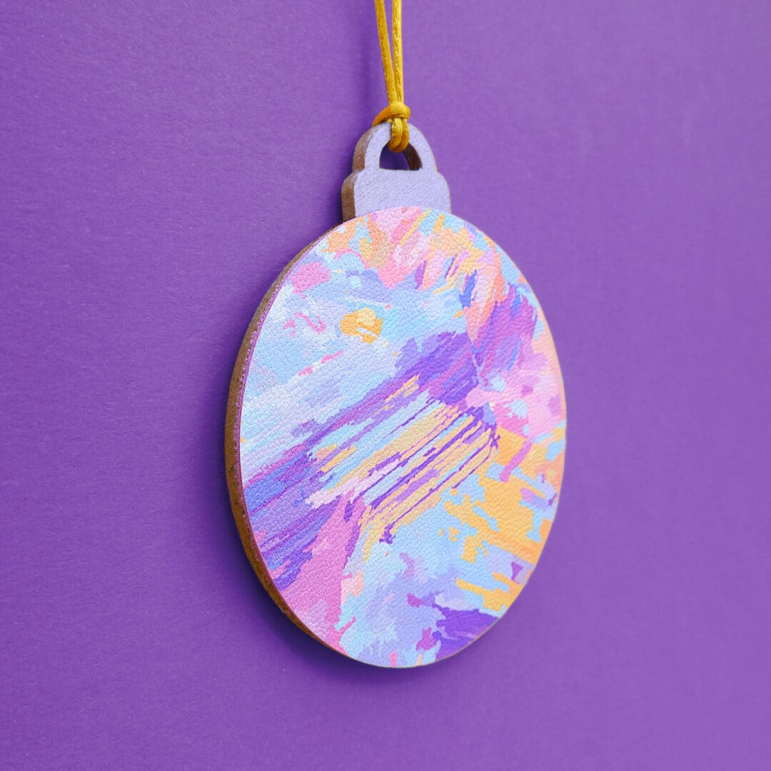 2D wooden Christmas bauble. Painterly abstract pattern in colours of purple, pink, orange, and blue. 7cm diameter wood and faux leather