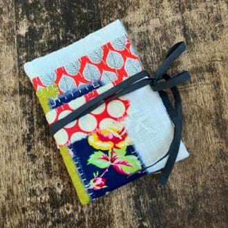 The photo shows a small needle book made of white embroidered fabric sitting on a battered wooden table top. The book is decorated with colourful fabric in contemporary designs, hand stitched onto the white main fabric. The needle book is shown fastened by grey ribbon which wraps around the book and ties with a bow.