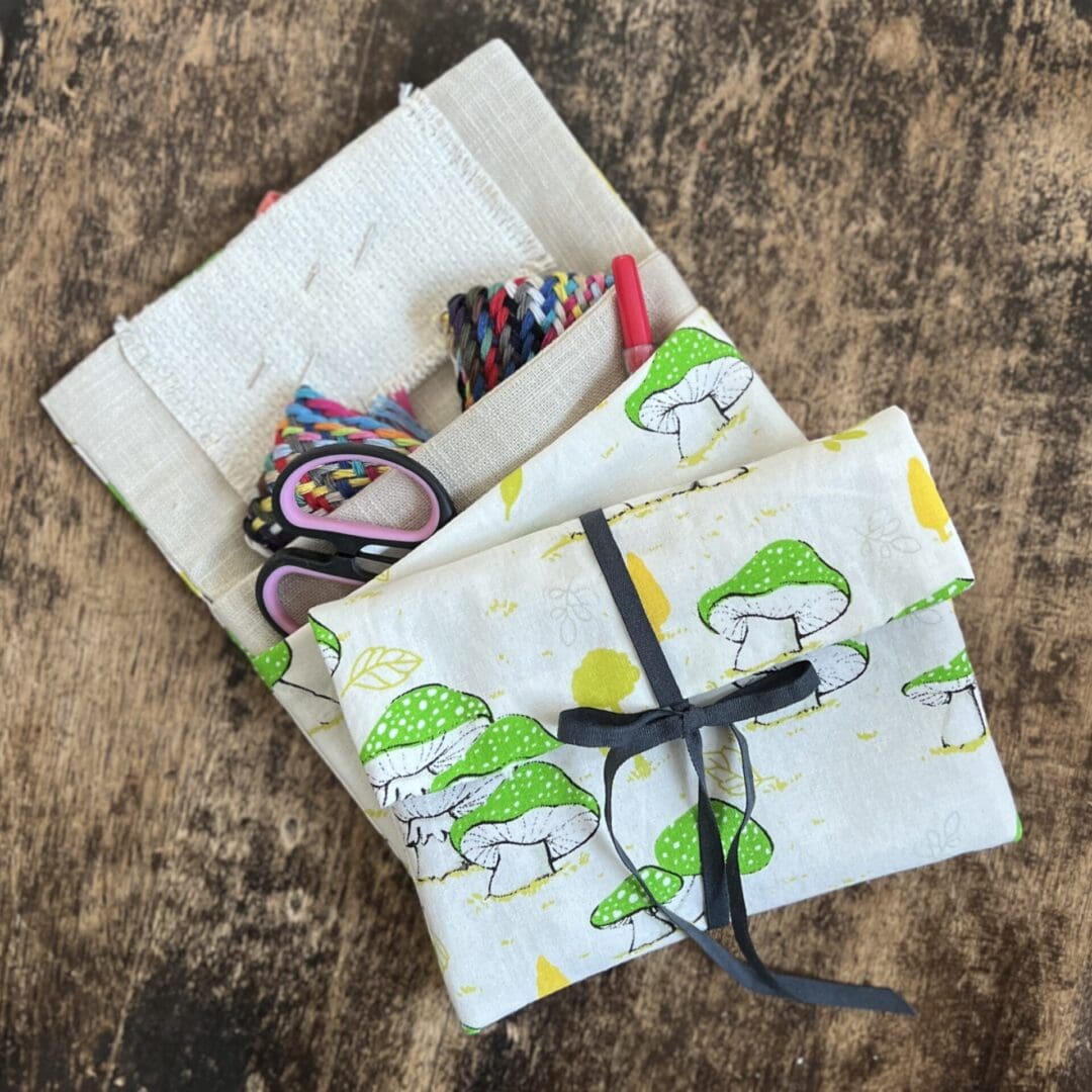 Two small cotton fabric pouches lay on top of each other on a wooden table top. Both pouches are made from a white cotton fabric with a green mushroom pattern. The pouches lye one atop the other, the top one fastened with a grey ribbon and ties in a bow. The other case is open showing its two internal pockets and the contents - a pair of scissors, a quick unpick and a multi coloured thread plait.