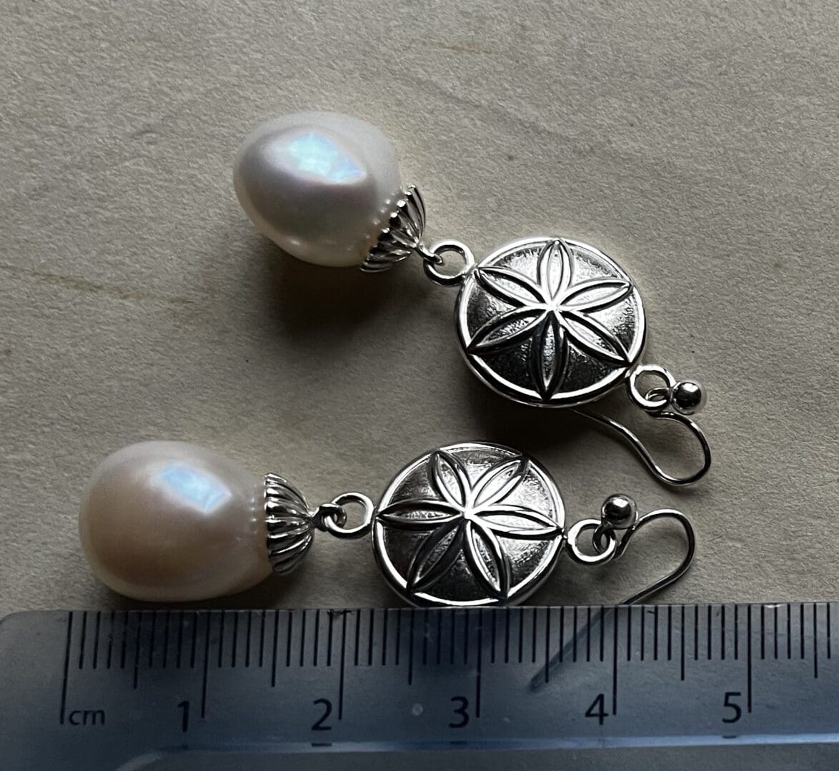 a pair of earrings with a silver daisy wheel symbol and a baroque pearl underneath it. There is a ruler next to the earrings stating the total drop length that says 40mm.