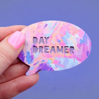 White woman's hand holding a handmade 'Day Dreamer' speech bubble brooch with a whimsical abstract design in pink, lilac, and yellow.