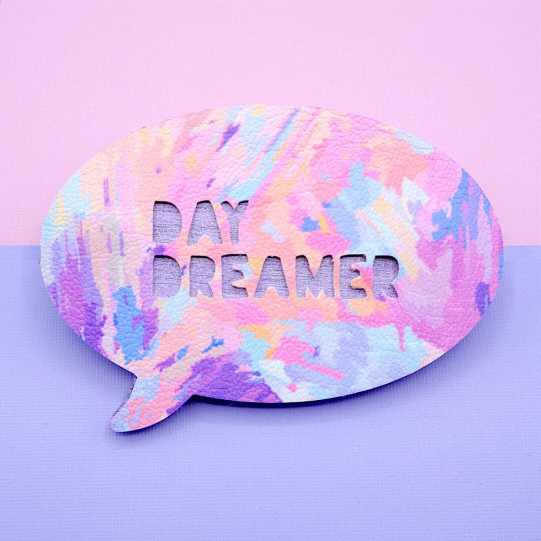 Handmade 'Day Dreamer' speech bubble brooch with a colourful painted abstract design in pink, lilac, and yellow, displayed on a pastel pink and lilac background