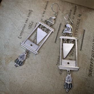 A pair of guillotine earrings in silver. There is a phrygian cap on top of the frame, a silver blade is suspended by small loops and underneath the frame is a head upside down