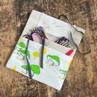 Picture shows a green mushroom patterned fabric pouch lying open on a wooden table top. The pouch has two pockets with scissors, quick unpick and a thread plait pocking out of them.