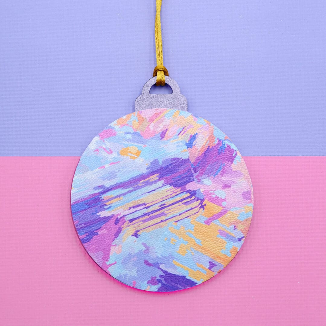 Handmade Christmas bauble with a vibrant abstract design in pink, lilac, yellow, and blue, displayed on a pastel pink and lilac background.