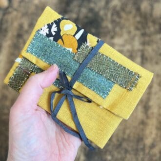 Picture shows a mustard linen fabric case decorated with a hand stitched patchwork of green woollen fabrics. The pouch lays across the makers hand with a wooden table top as the background.