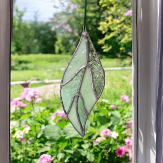 Handmade stained glass leaf suncatcher in light green, hanging by a green ribbon in a sunny window. Adds spring decor to the home; perfect gift for nature lovers.