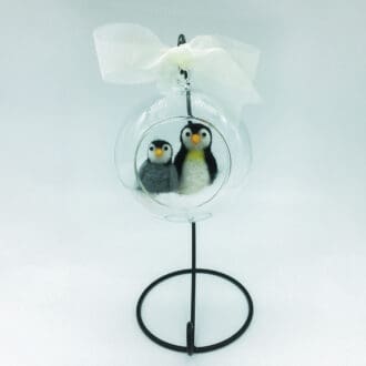 Full view of a handcrafted needle-felted adult penguin and chick inside a glass globe with a white bow, displayed on a sturdy black metal stand for holiday displays.