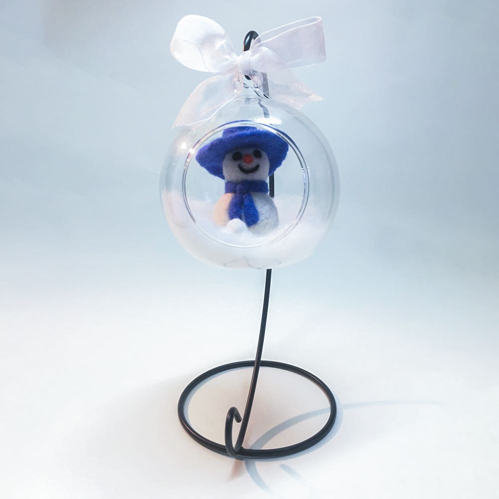 Full view of a needle-felted snowman bauble in a glass globe with soft wool snow, displayed on a black metal stand for holiday decor.