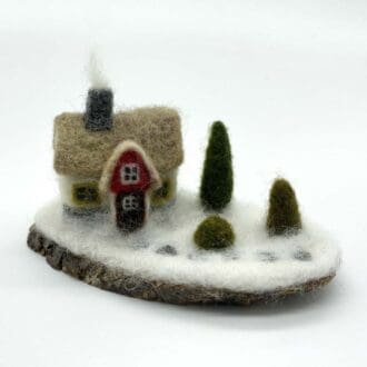 Front view of a handcrafted needle-felted winter cottage sculpture featuring a red door, chimney with smoke, and snow-covered base on a rustic wooden platform.