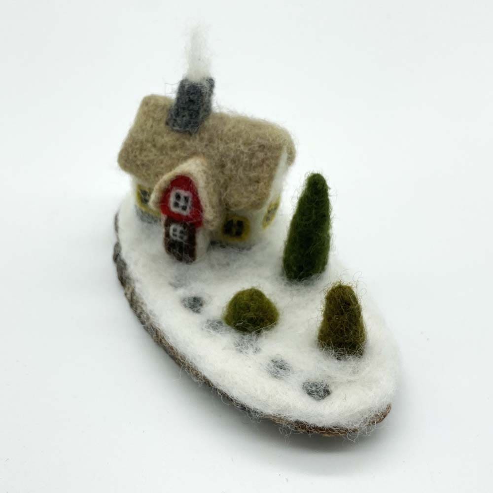 Top view of a needle-felted winter cottage sculpture, showcasing the chimney, roof, trees, and snow-covered details on a wooden base