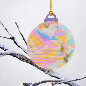 Pastel Christmas bauble with abstract yellow, pink, and blue design, hanging from a gold string on a snowy branch, handmade from wood and fabric