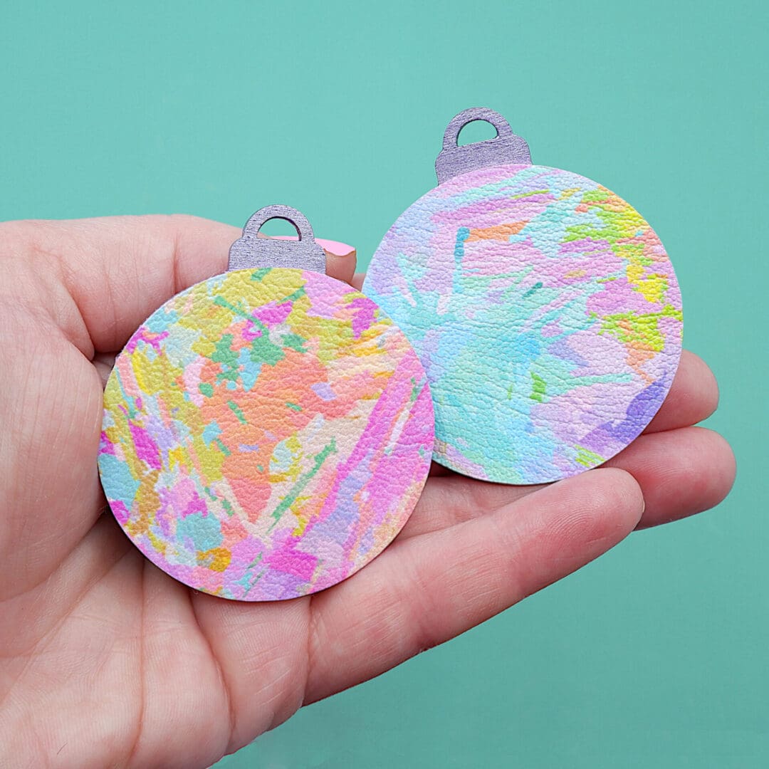 Two flat, round Christmas baubles with pastel printed abstract art. Carries in a white woman's hand.