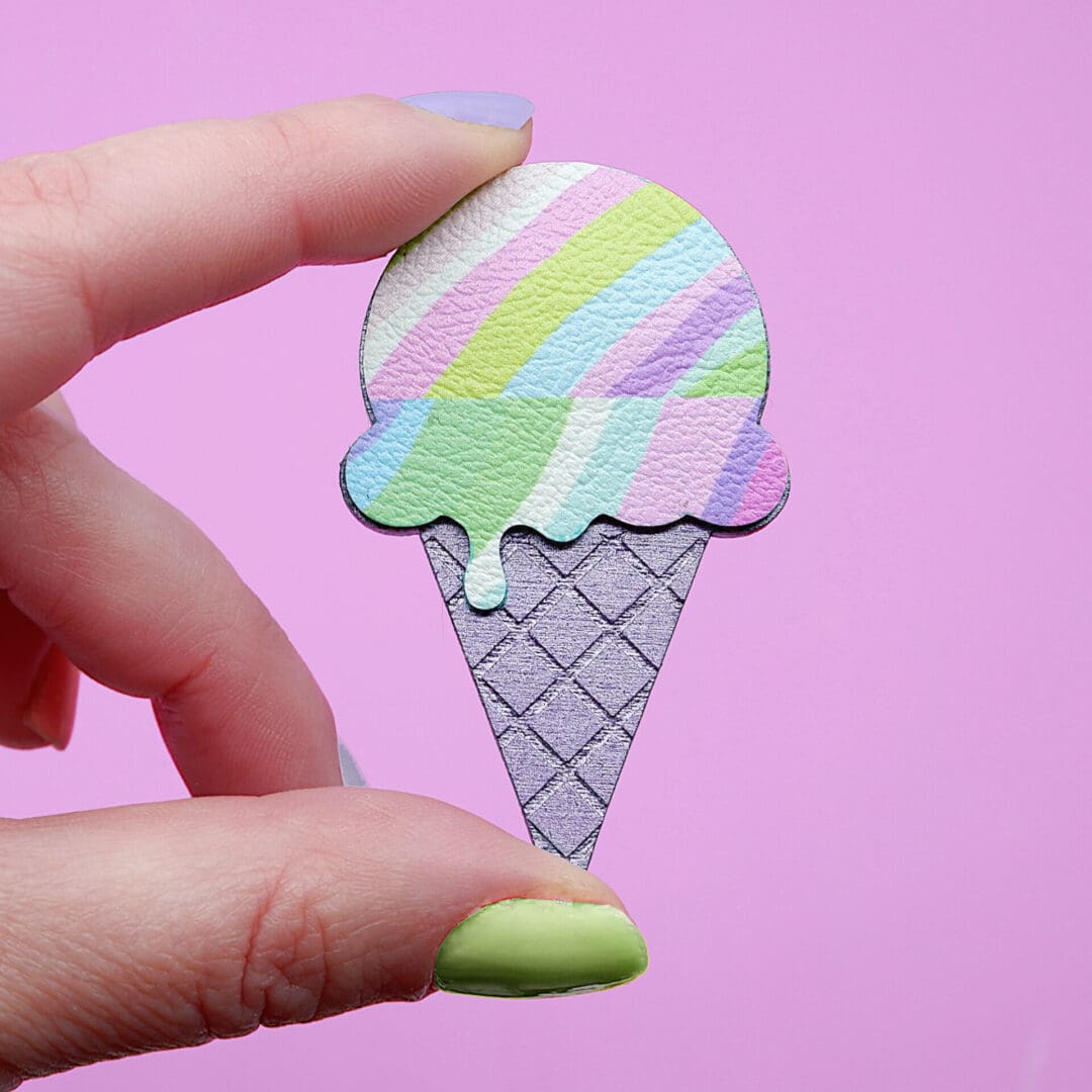 Pastel ice cream-shaped brooch with colourful stripes and a waffle cone design, handmade with a playful and whimsical aesthetic, shown against a pink background.