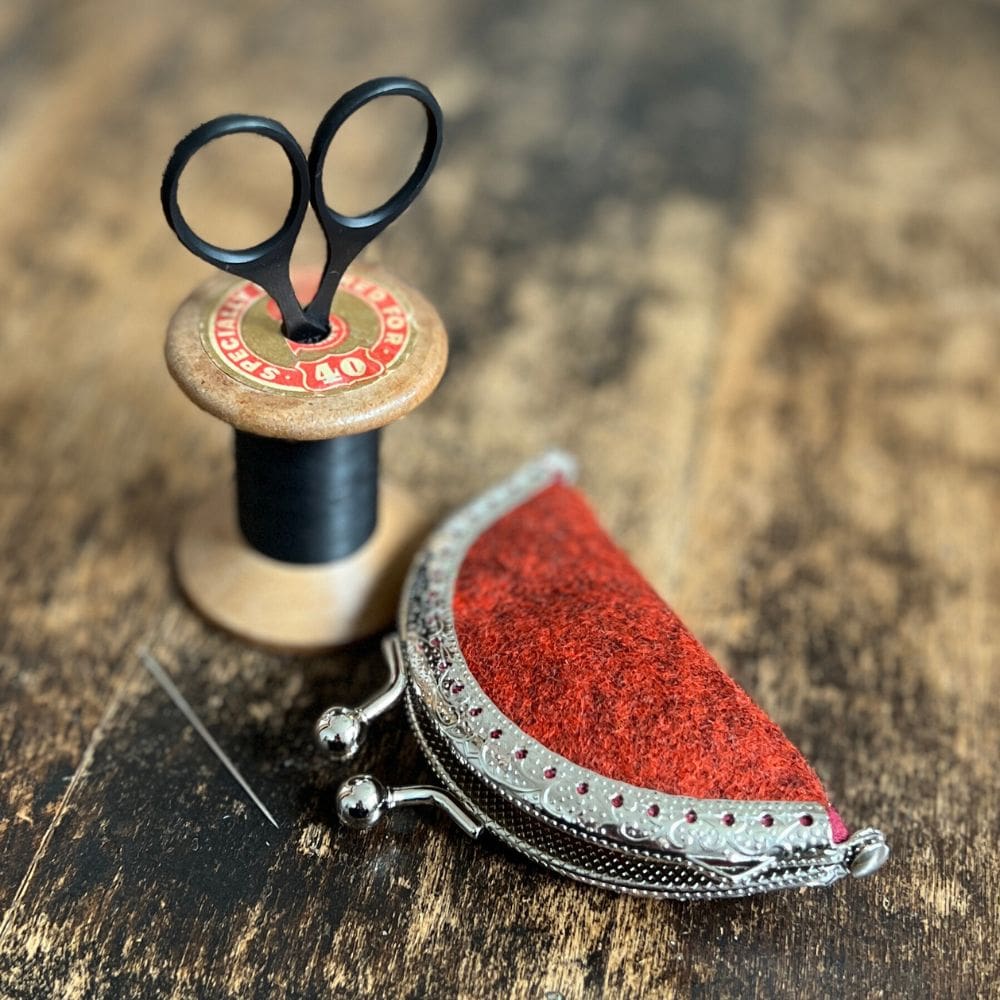 A small, metal clasp fastening needle book, made of burnt orange wool fabric, lays on a wooden table top. Next to the needle case is a vintage cotton thread bobbin with small, black scissors.