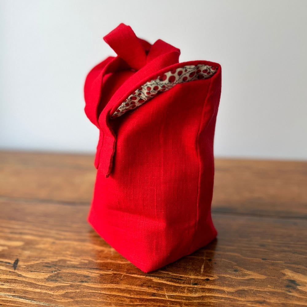 The picture shows a bag made of red linen sitting atop a wooden table top. The bag is sideways on allowing a peek at the cream and red patterned lining showing in the slightly turned bag top flap.