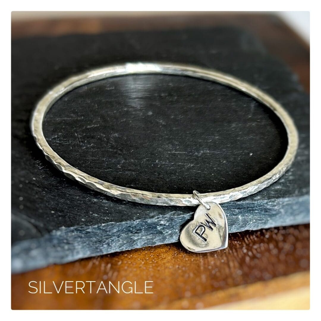 A Hammered Silver Bangle with a silver heart charm hand stamped with tow capital letters sits on a grey slate