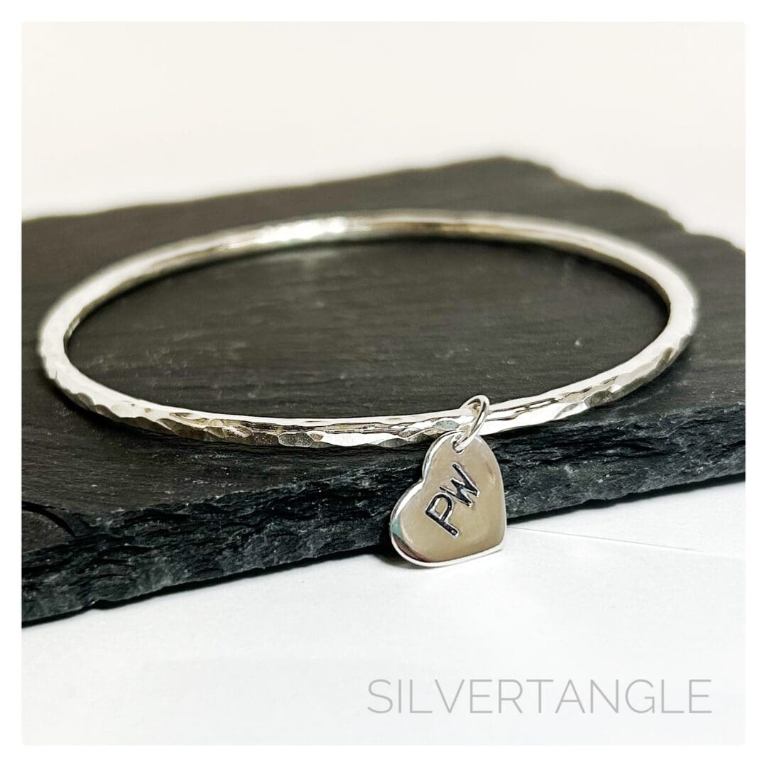 A Hammered Silver Bangle with a silver heart charm hand stamped with tow capital letters sits on a grey slate