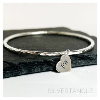 A Hammered Silver Bangle with a silver heart charm hand stamped with tow capital letters sits on a grey slate