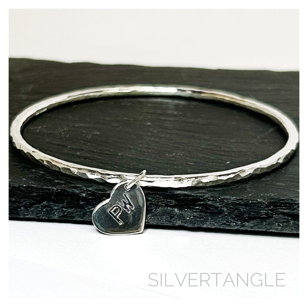 A Hammered Silver Bangle with a silver heart charm hand stamped with tow capital letters sits on a grey slate