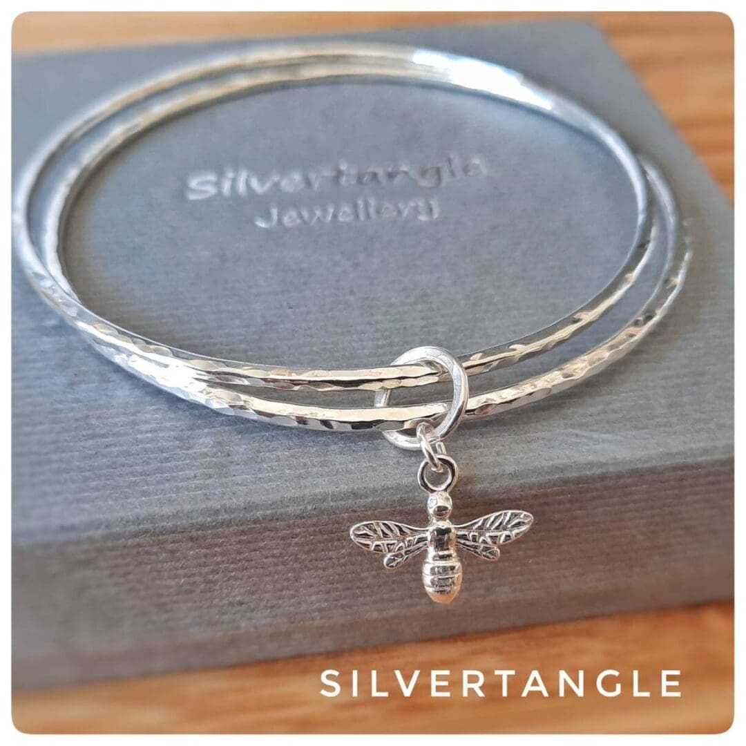 two silver hammered bangles with a bee charm sitting on a grey box