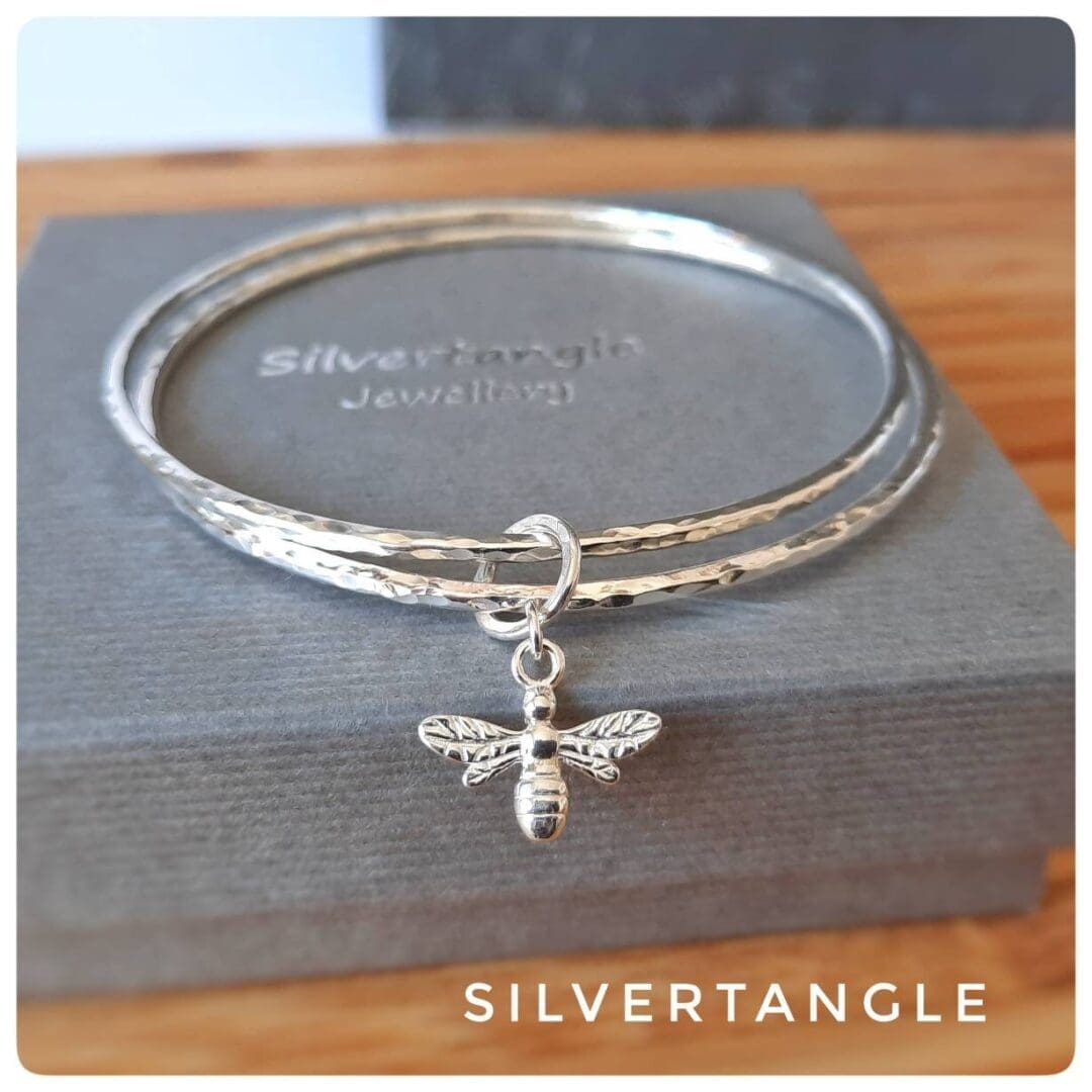 two silver hammered bangles with a bee charm sitting on a grey box