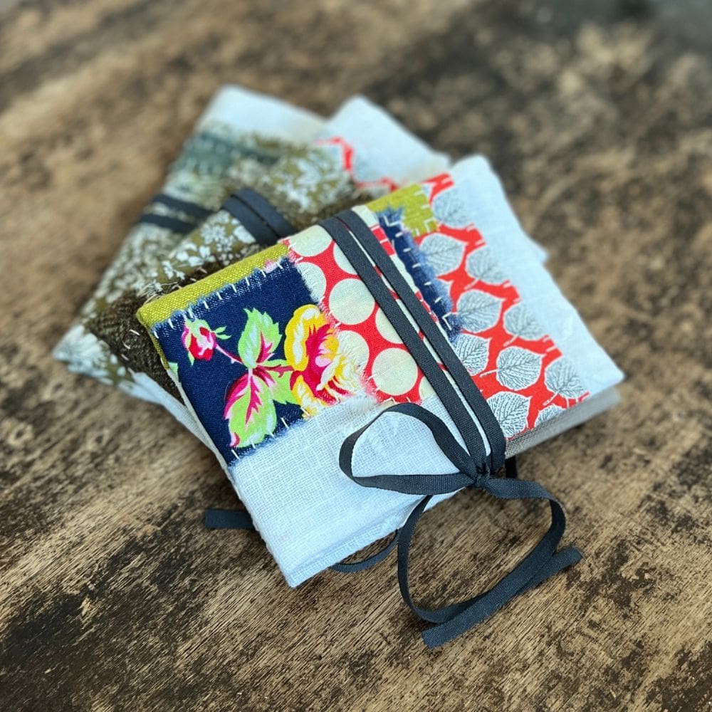 A collection of three patchwork needle books sit on a battered wooden table top. The books are made from a white embroidered cotton, with patches of coloured fabrics of a contemporary design hand stitched to the main fabric. The books fasten with a grey ribbon which is wrapped around the book and tied with a bow.