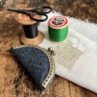 A petite, blue wool needle book with a metal purse clasp fastener sits on a wooden top surrounded by vintage haberdashery