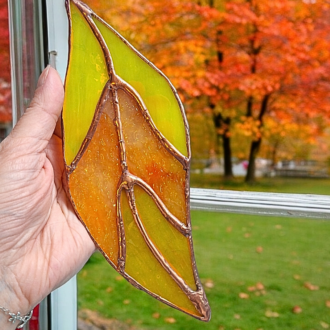 Bring the beauty of autumn indoors with this stunning handmade stained glass leaf! Measuring 19 cm long and 7 cm at its widest, this nature-inspired glass art features rich autumn colours, a glowing copper patina, and hangs from a purple ribbon. Perfect for rustic decor and plant lovers seeking unique seasonal decor.