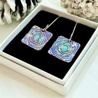 À pair of copper drop earrings in a square shape with a intricate pattern and hand painted in blue and Green metallic paints, they have sterling silver ear wires in a fish book shape. The earrings are displayed on a black velvet insert in a White gift box.